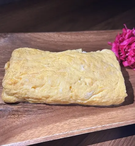黄色く焼き上げただし巻き玉子焼き。木の板に紅色の漬物を添えて